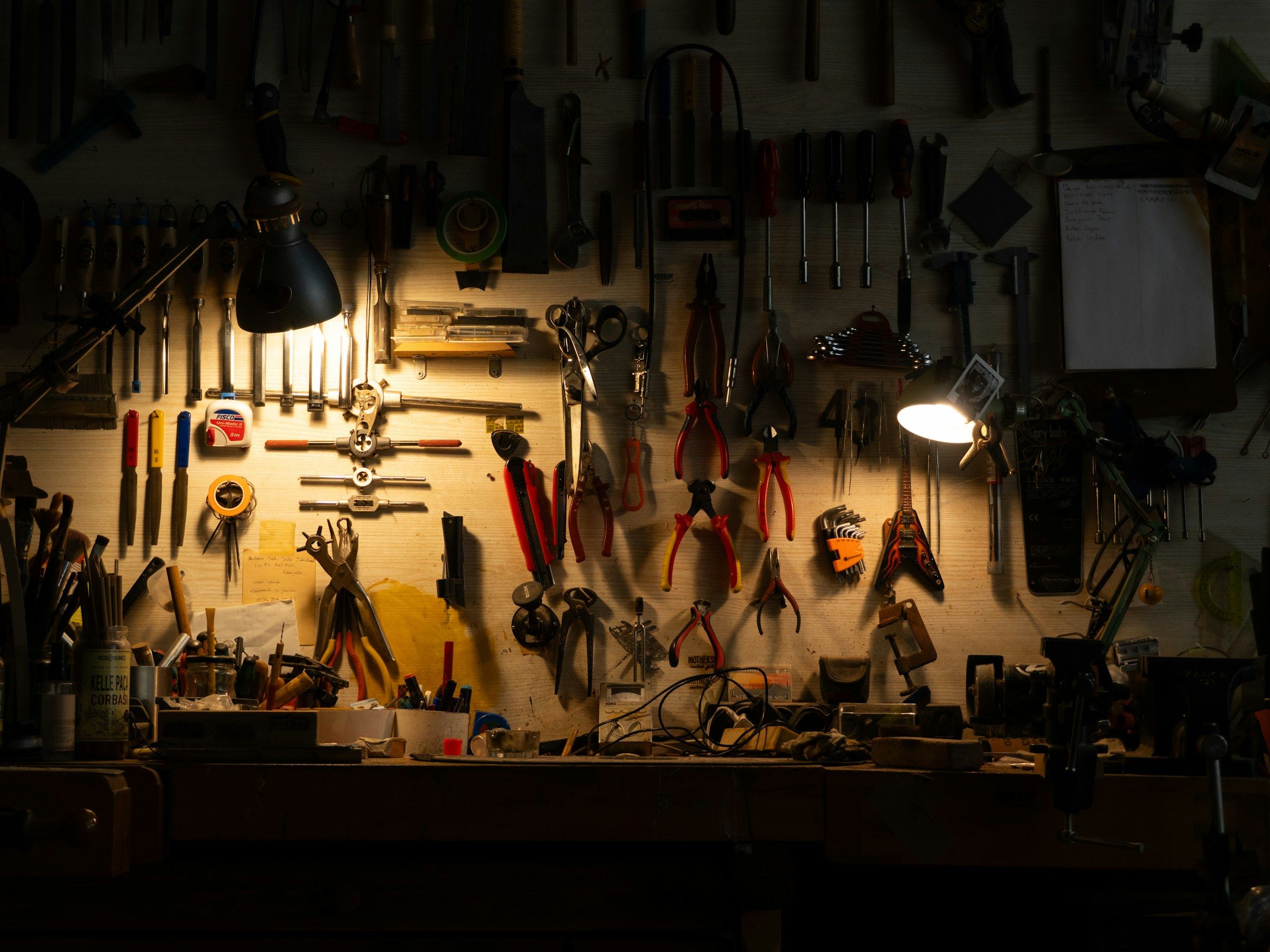 Workbench wall of hanging hand tools lit by two lamps in a dark workshop