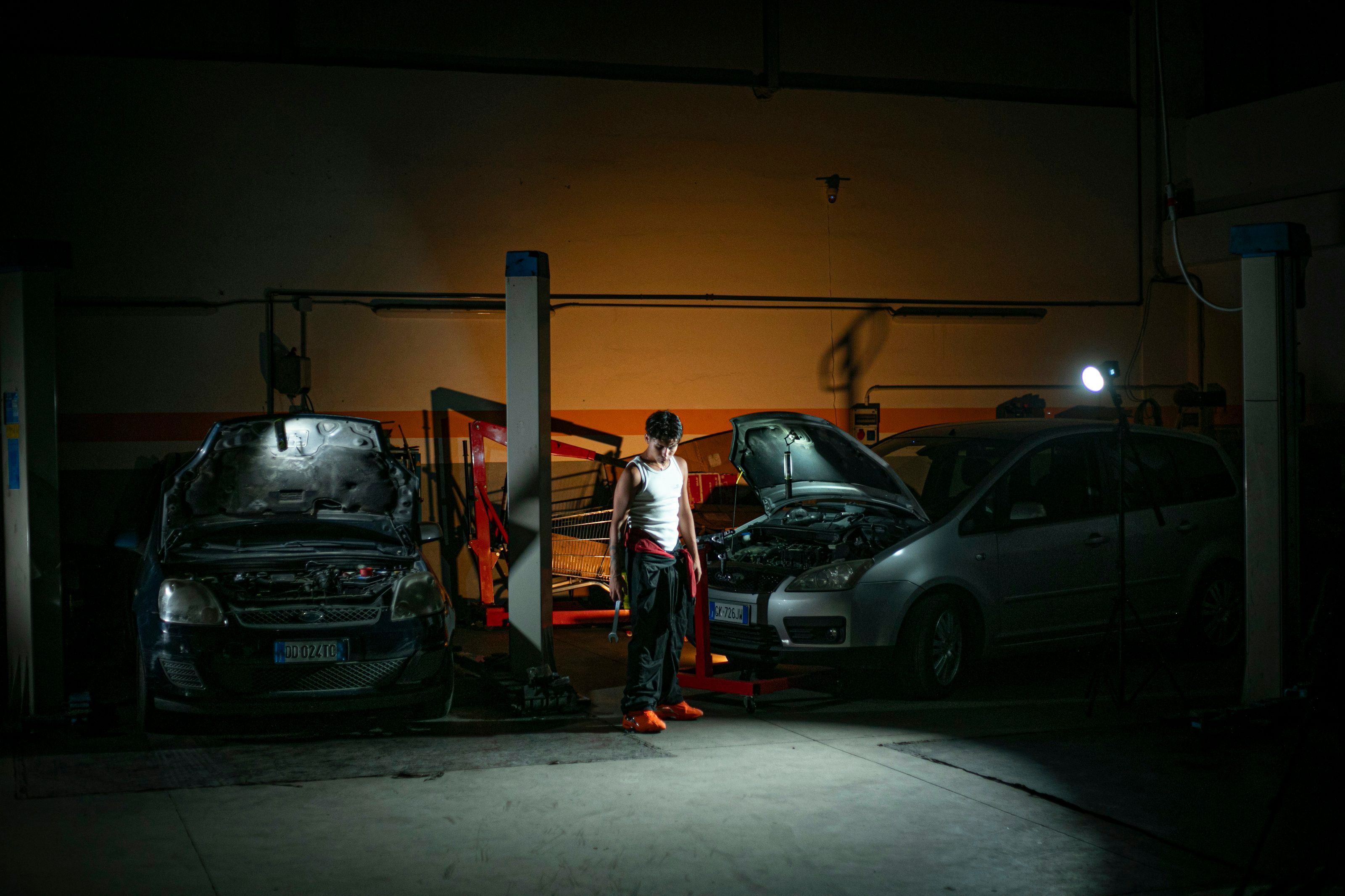 Two cars on lifts with their hoods up in a dark garage at night, a mechanic standing under a single work light