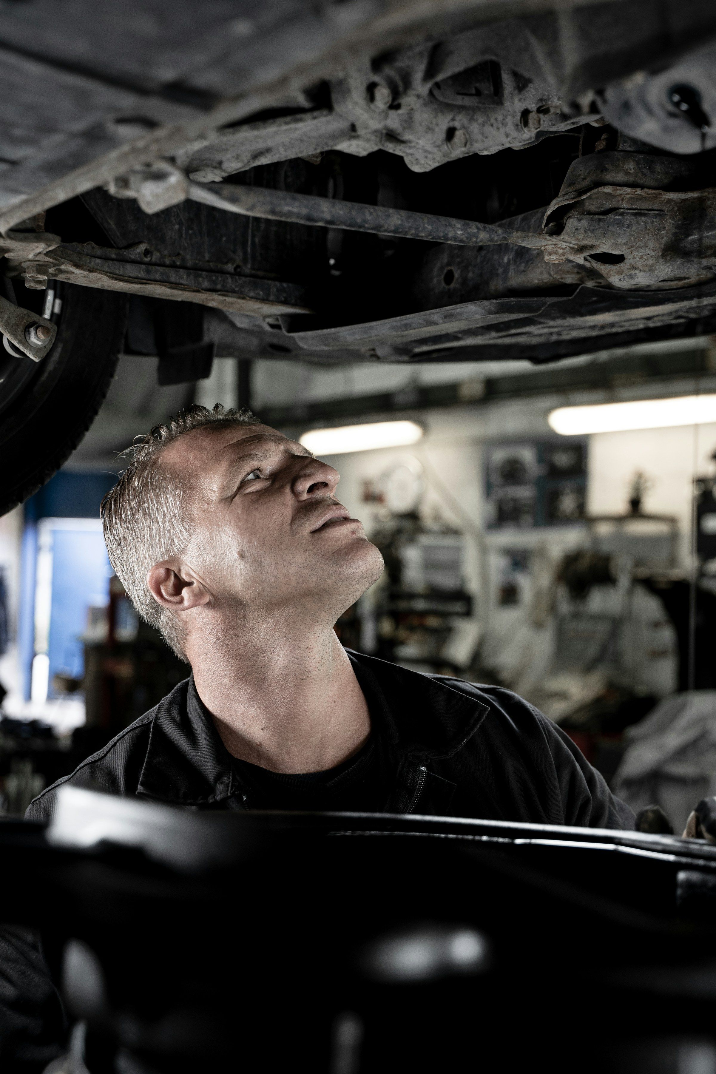 Gray-haired mechanic standing under a lifted car, looking up at the underside
