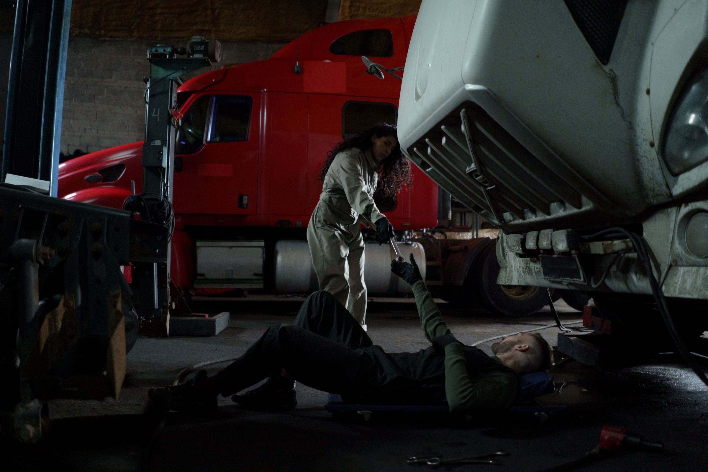 Two mechanics repairing a white truck in a dim garage, one lying on the floor beneath it while the other works at the front