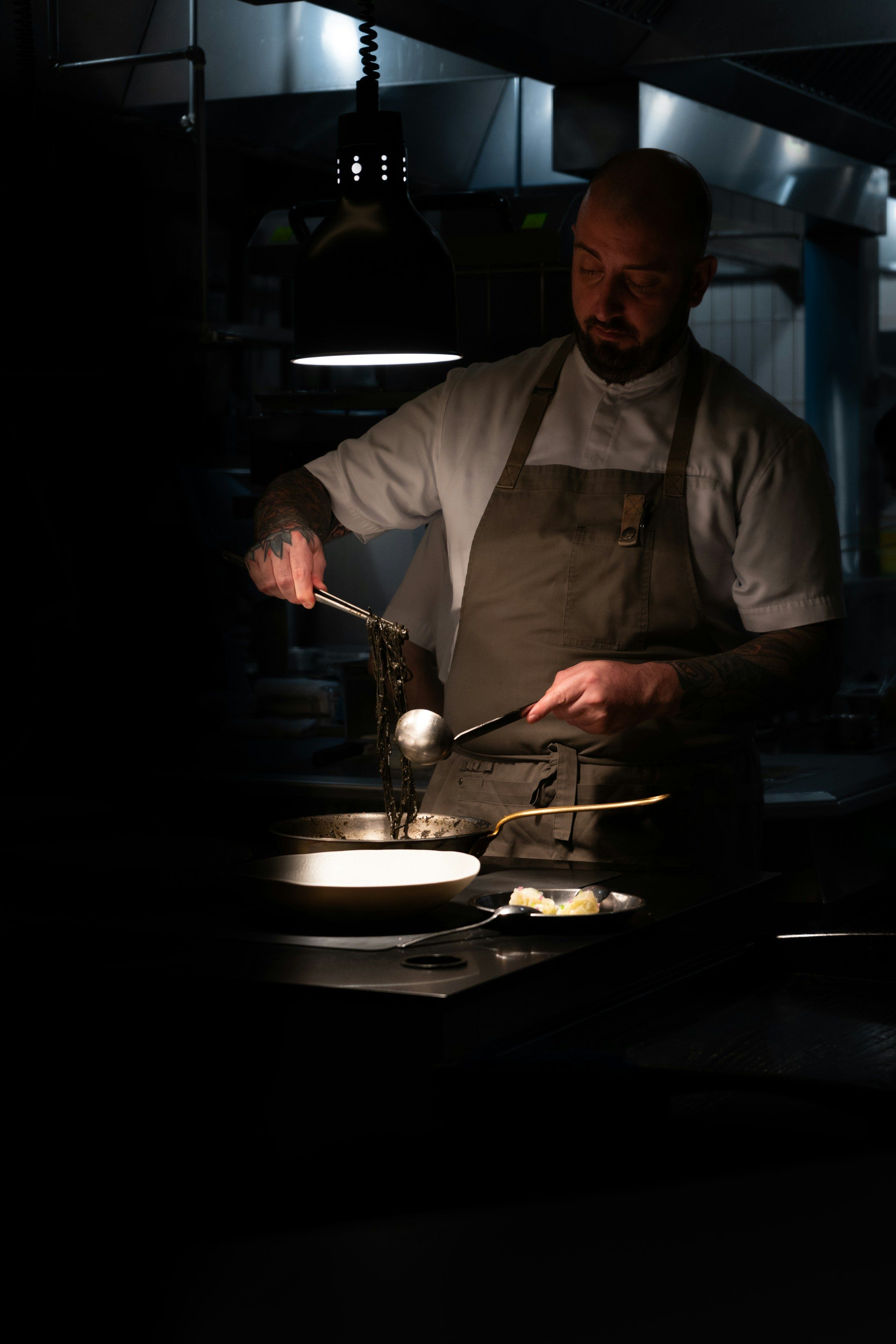 A chef lifts food from a pan with tongs under a heat lamp in a dark kitchen.