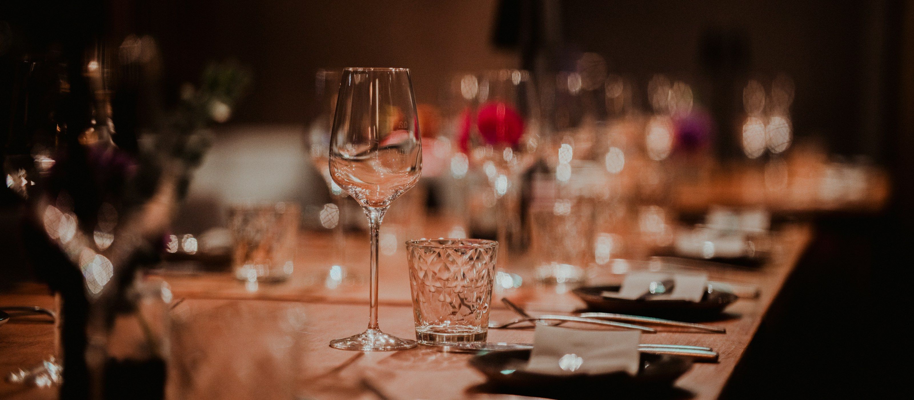 A long table set with wine glasses, tumblers and folded napkins, softly blurred in warm evening light.