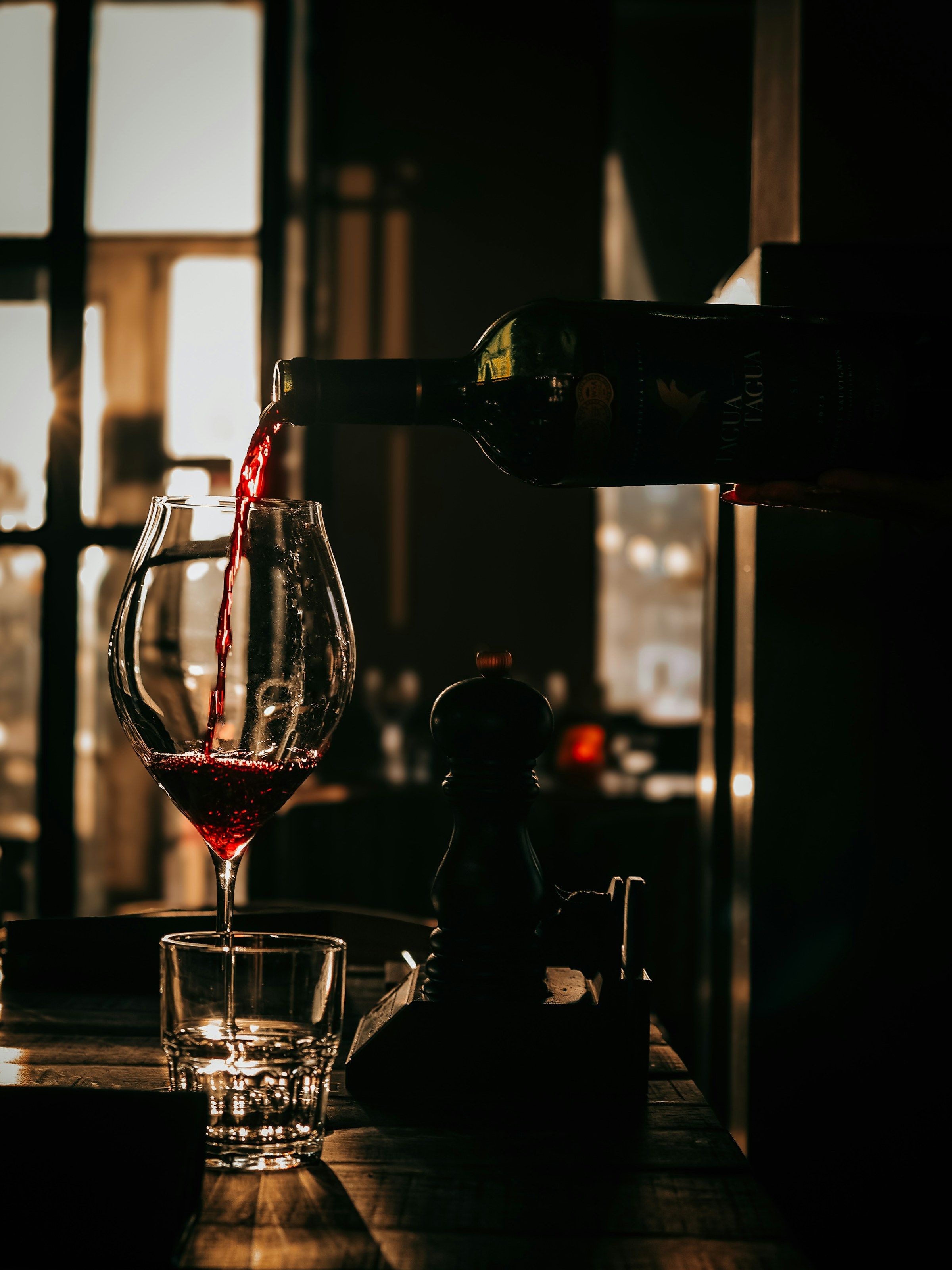 Red wine pouring into a glass on a dark table, backlit by a window.