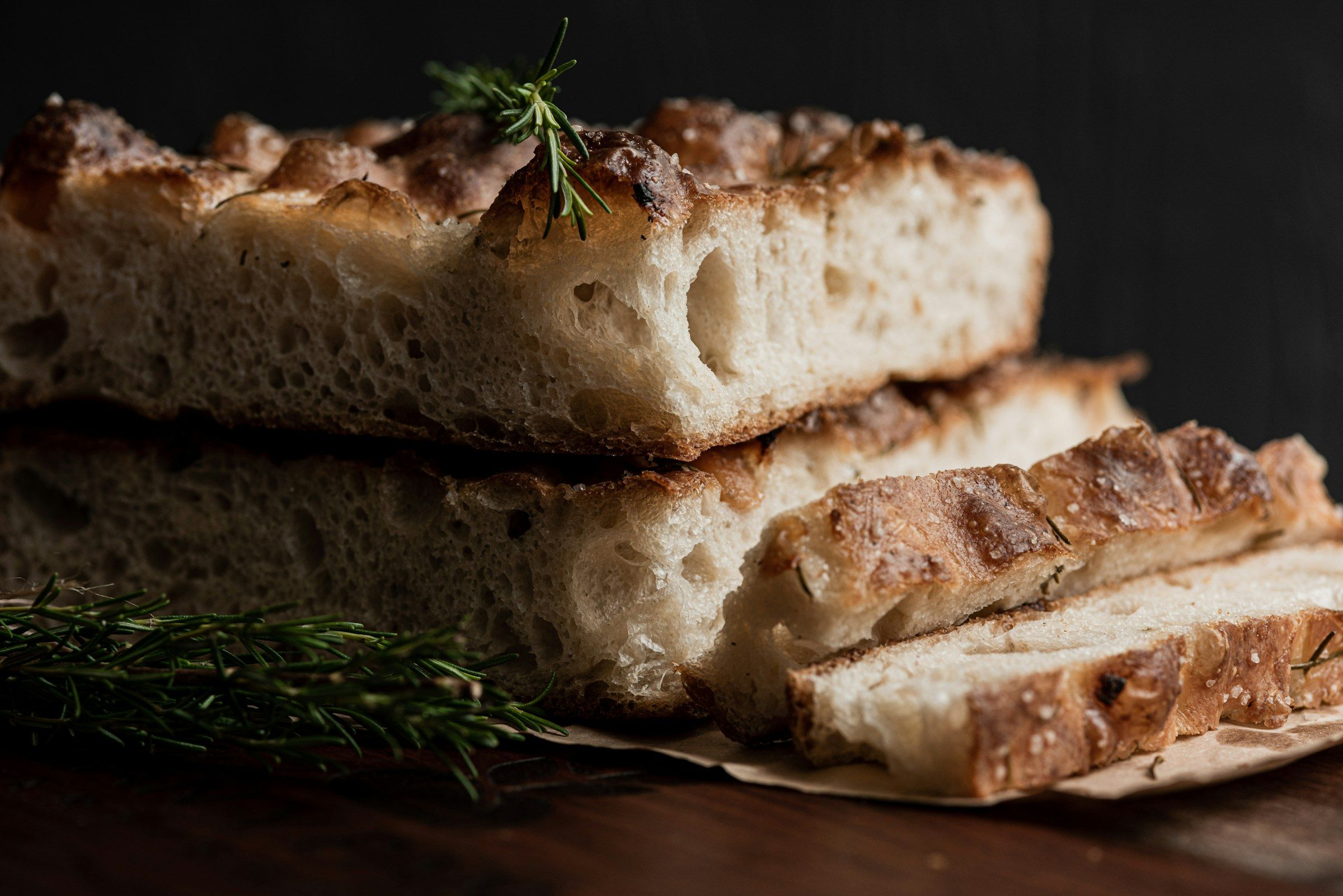 Slices of golden focaccia with rosemary stacked on parchment against a dark background.