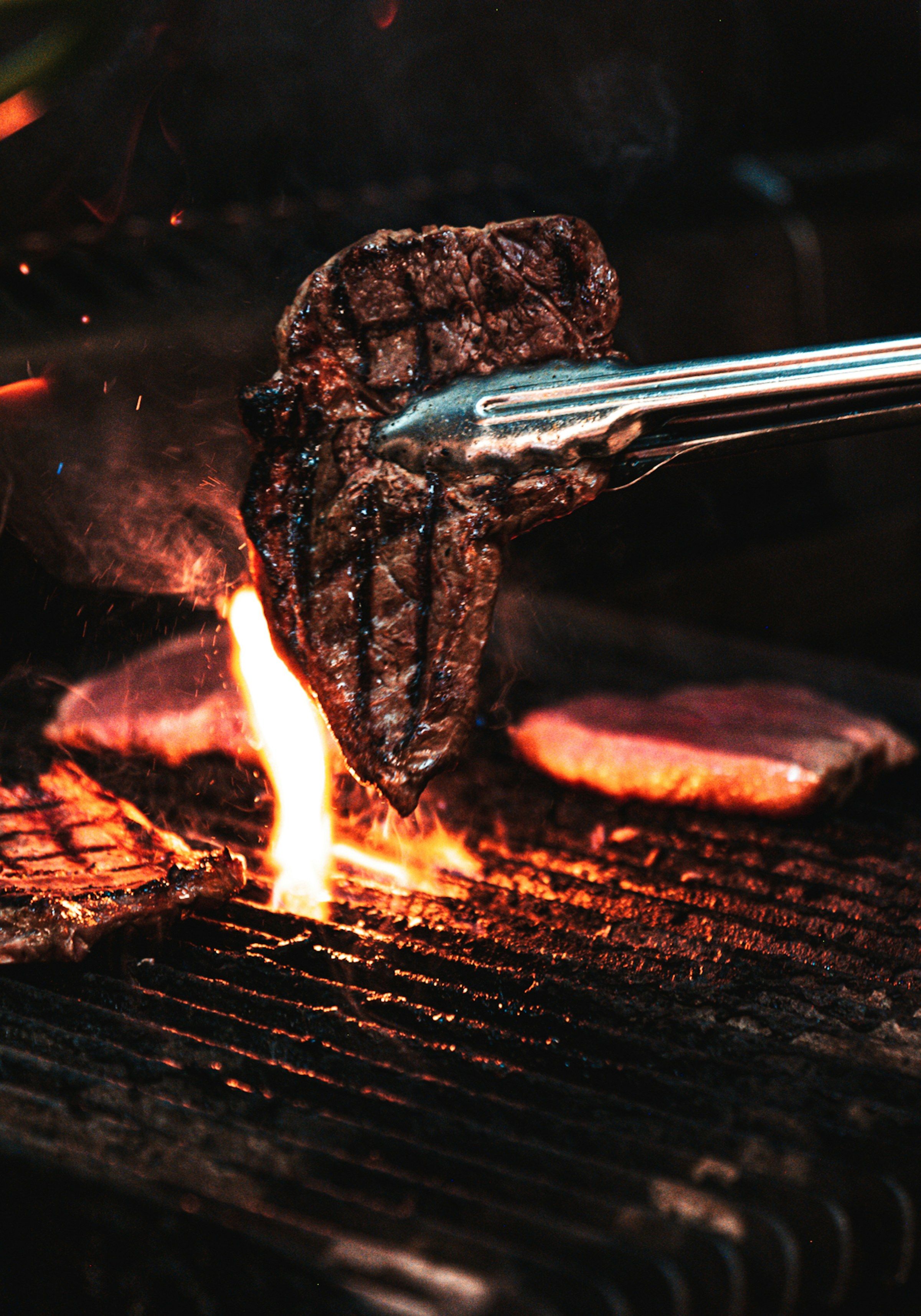 Tongs lifting a seared steak over the flames of a charcoal grill.