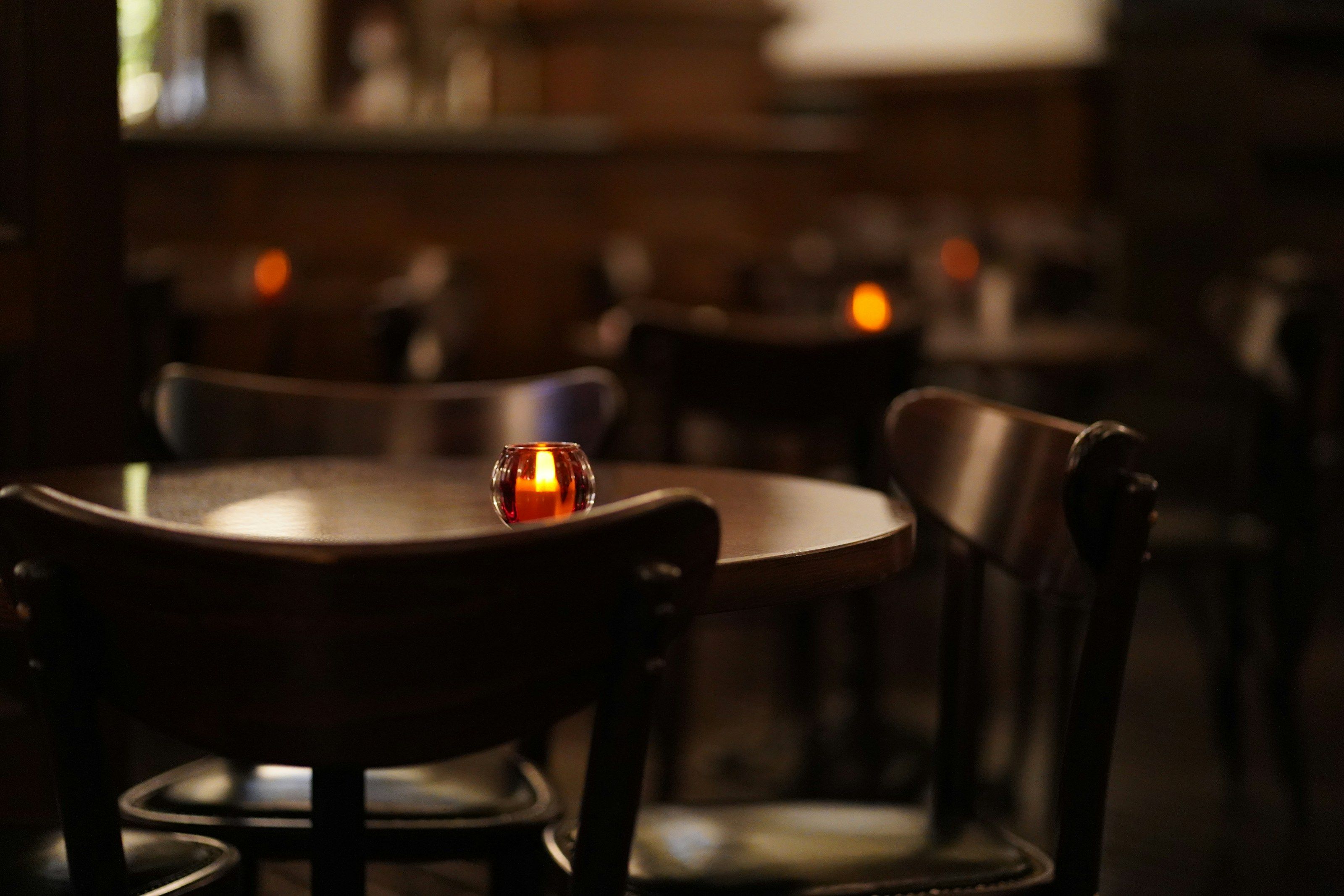 A small candle glows on a dark wooden bistro table between bentwood chairs in a dimly lit dining room.