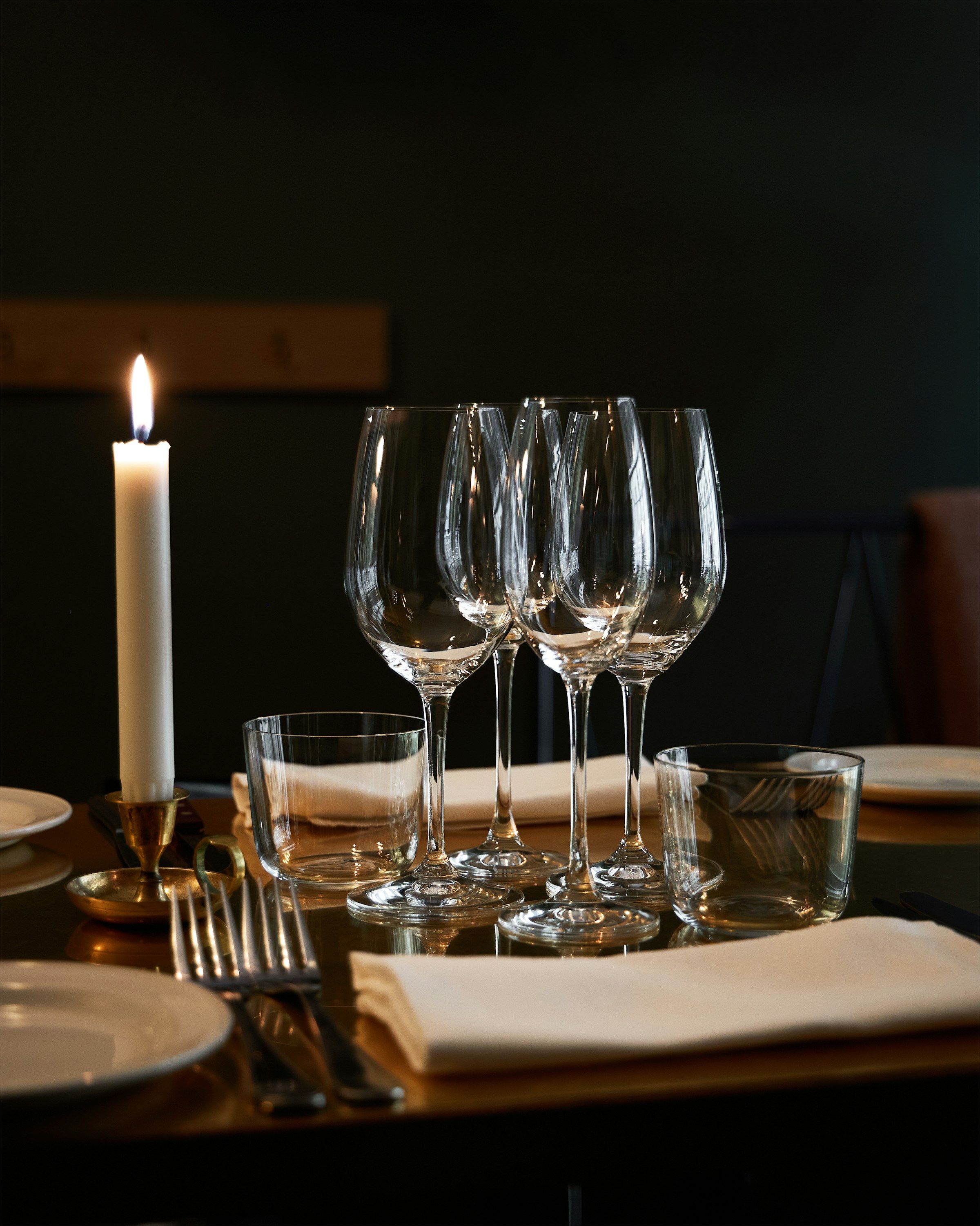 A lit taper candle beside empty wine glasses and folded napkins on a set table in low light.