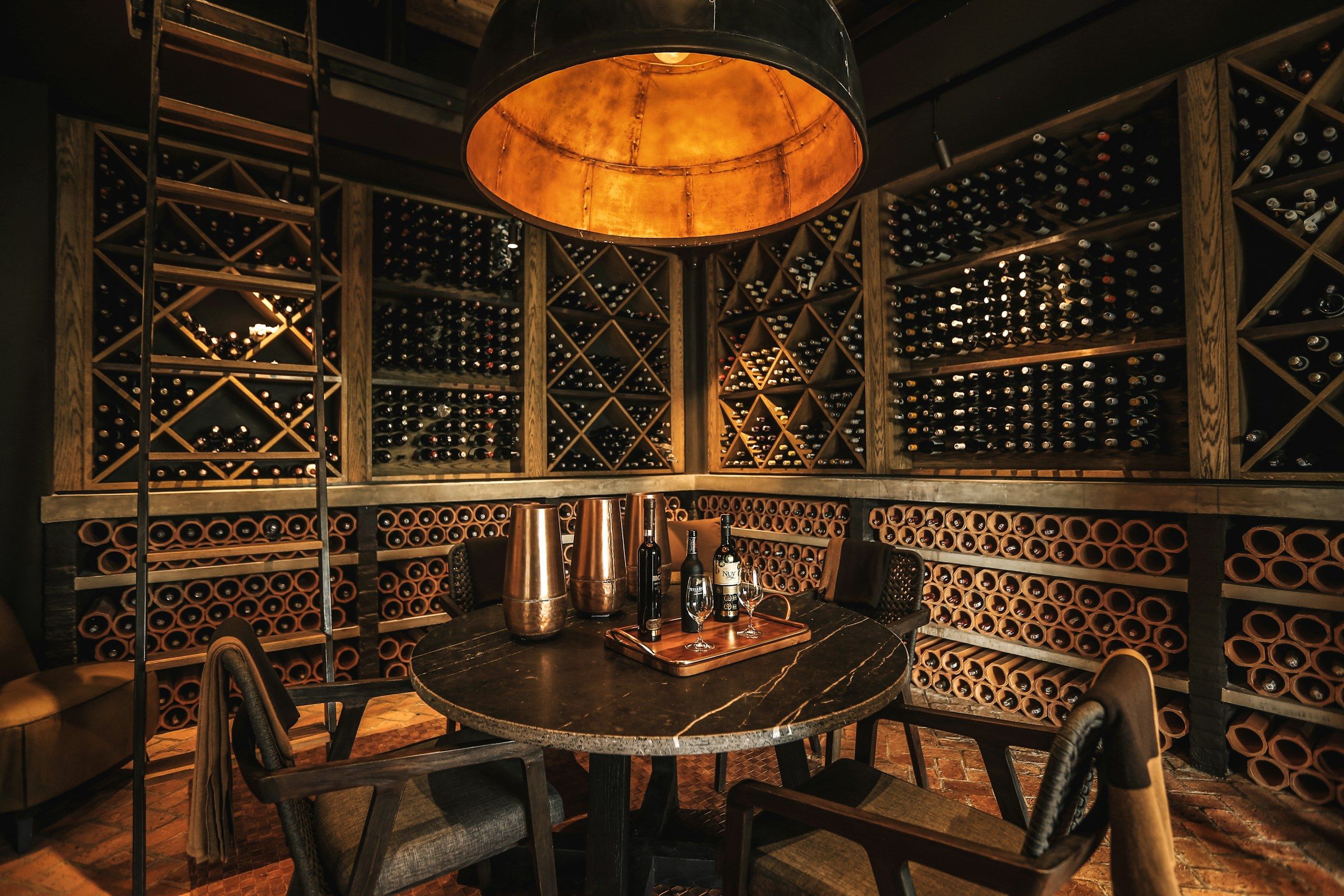 A round table in a wine-cellar dining room, surrounded by bottle racks under a copper pendant lamp.