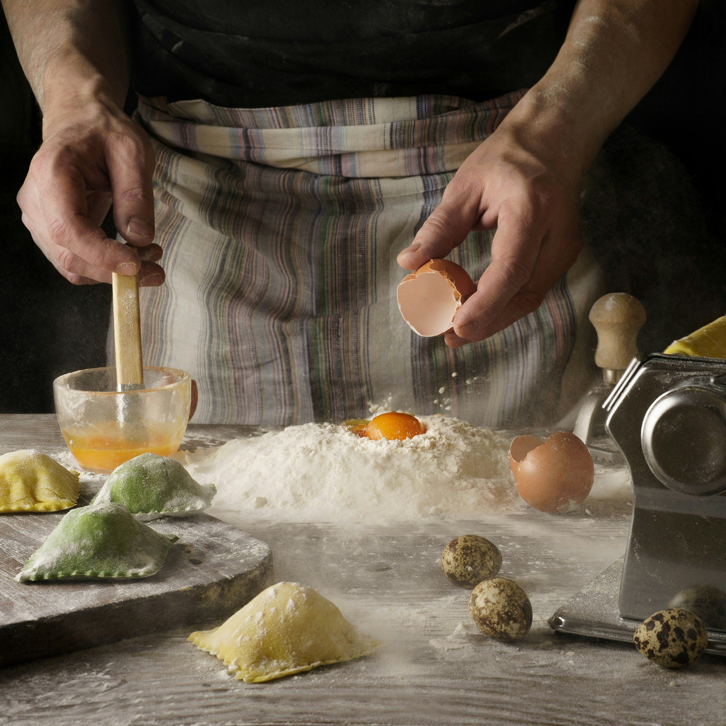 Hands cracking an egg beside a well of flour and yolk, with fresh ravioli, eggs and a pasta machine on the board.